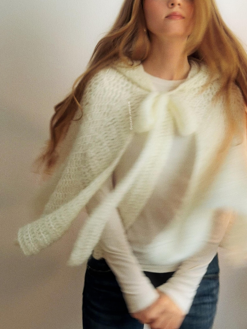 May include: A cream-colored, hooded cape with a bow detail. The cape is made of a soft, open-weave knit fabric. The image shows the cape in motion, with the fabric flowing. The cape is worn over a white long-sleeved shirt and blue jeans.