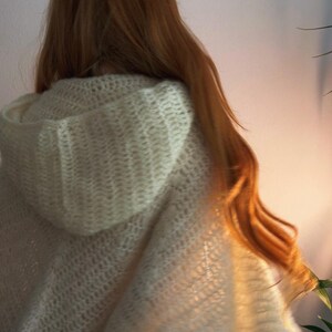 May include: Cream-colored hooded cape with a textured, open-knit design. The cape is draped over the shoulders, with long, flowing auburn hair visible. The hood is up, and the fabric appears soft and cozy.