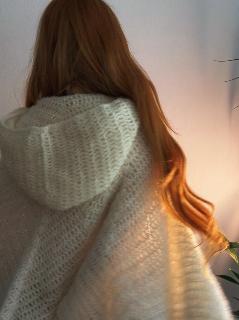May include: Cream-colored hooded cape with a textured, open-knit design. The cape is draped over the shoulders, with long, flowing auburn hair visible. The hood is up, and the fabric appears soft and cozy.