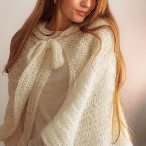 May include: A cream-colored, open-weave crochet cape with a hood and a bow closure. The cape is made of a soft, textured yarn and drapes over the shoulders. The overall style is bohemian and feminine.