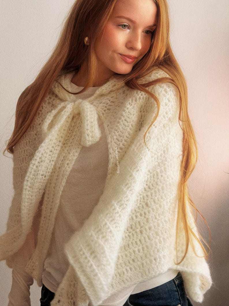 May include: A cream-colored, open-weave crochet cape with a hood and a bow closure. The cape is made of a soft, textured yarn and drapes over the shoulders. The overall style is bohemian and feminine.