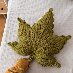 Può includere: Un cuscino a forma di foglia all'uncinetto, verde oliva fatto a mano. Il cuscino ha la forma di una grande foglia con venature dettagliate e una superficie strutturata. Una persona con un maglione giallo senape tiene il cuscino.