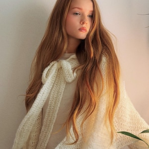 May include: A cream-colored, open-weave shawl with a bow detail. The shawl is draped over a person, with long auburn hair cascading down. The garment appears soft and lightweight, suitable for layering. The background is a plain, light-colored wall.