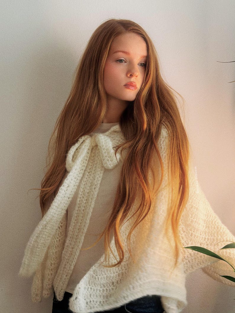 May include: A cream-colored, open-weave shawl with a bow detail. The shawl is draped over a person, with long auburn hair cascading down. The garment appears soft and lightweight, suitable for layering. The background is a plain, light-colored wall.