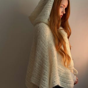 May include: A cream-colored, hooded poncho with a diamond-patterned knit design. The poncho is made of a soft, textured material and drapes over the shoulders. The hood is large and covers the head. The garment is worn over a white shirt and blue jeans.