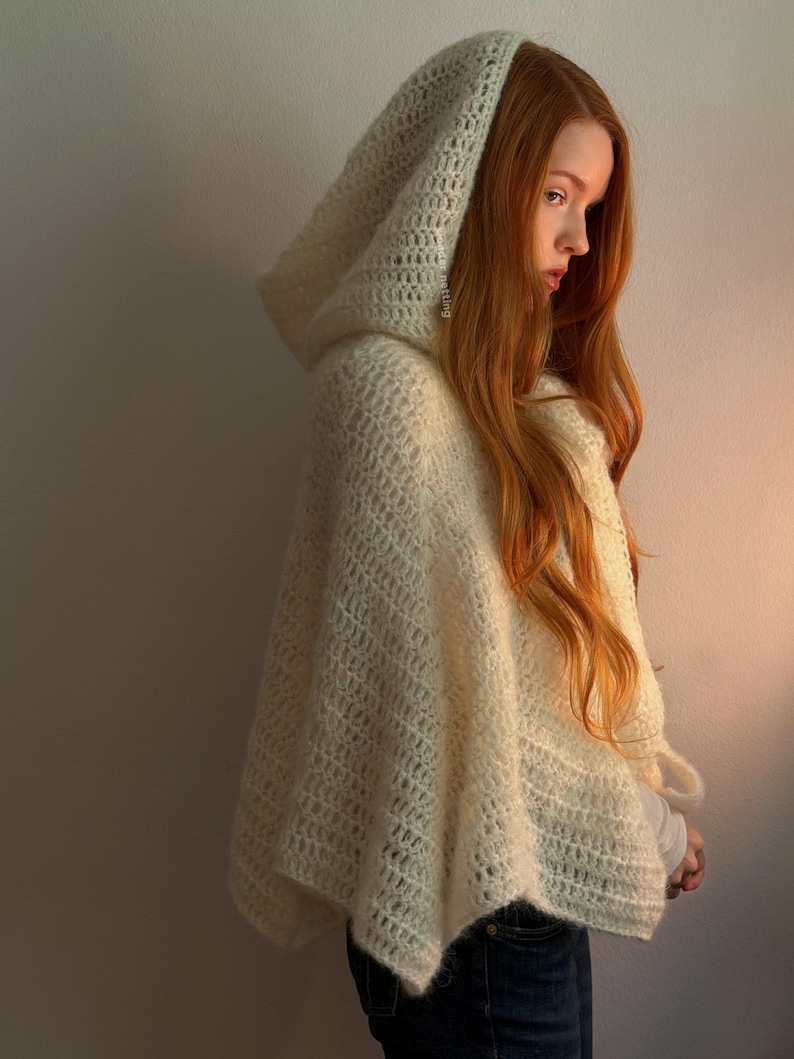 May include: A cream-colored, hooded poncho with a diamond-patterned knit design. The poncho is made of a soft, textured material and drapes over the shoulders. The hood is large and covers the head. The garment is worn over a white shirt and blue jeans.