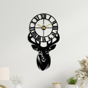 Puede incluir: Reloj de pared de metal negro con un diseño de cabeza de ciervo y números romanos. El reloj está colgado en una pared blanca sobre una cómoda de madera.