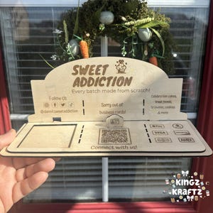 Peut inclure: Un présentoir en bois avec le texte "SWEET ADDICTION" et "Every batch made from scratch!". Il comprend des identifiants de réseaux sociaux, un code QR et des options de paiement. Le présentoir est tenu devant une fenêtre avec une couronne décorative.