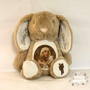 May include: A plush brown bunny rabbit with a centered image of a woman. The bunny's ears have the text "Billie Eilish" and "Happier Than Ever". The bunny has a white belly and a black silhouette of a person.