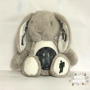 Puede incluir: Un conejo de peluche gris con orejas largas. El conejo tiene una imagen circular en el pecho y el logo del cantante en el pie. El texto en las orejas dice "Billie Eilish" y "Hit Me Hard and Soft".