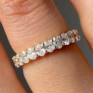 以下が含まれることがあります： 楕円形のダイヤモンドが多数セットされたゴールドバンドリング。 ダイヤモンドは密接に配置され、連続的な輝きを生み出しています。 リングは指に装着されており、そのデザインと輝きを強調しています。 リングのセッティングは、クラシックな爪のスタイルです。