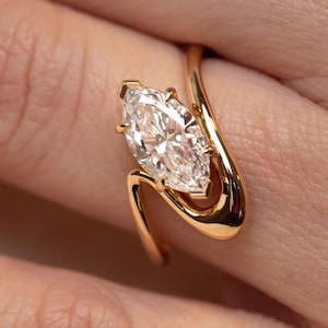 May include: A gold ring featuring a marquise-cut clear gemstone held in a prong setting. The band has a bypass design, with two curved ends that wrap around the central stone, creating an open, fluid silhouette on the finger.