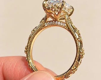 Puede incluir: Un anillo de oro con una gran piedra preciosa transparente, redonda y facetada, en un engaste de garras. La banda presenta detalles metálicos intrincados y elevados en forma de flores y hojas, con pequeñas piedras.