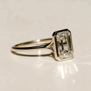 Op de afbeelding: Een gouden ring met een grote, rechthoekige, heldere edelsteen. De edelsteen is gezet in een bezel setting en de band is dun en gepolijst. De ring ligt op een wit oppervlak. De ring is een sieraad.