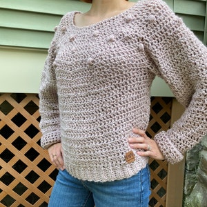 Puede incluir: Un suéter de punto de color rosa claro con un patrón de punto texturizado y una pequeña etiqueta de cuero marrón que dice "Love is Love".