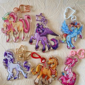 Puede incluir: Colección de llaveros acrílicos coloridos con personajes de dibujos animados de ponis. Cada llavero tiene un diseño diferente, con varios colores como rosa, morado, azul y amarillo. Los llaveros tienen cierres metálicos de varias formas.
