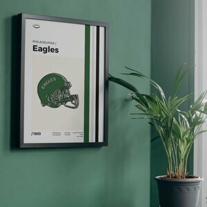 May include: A framed poster with a green and white striped background. The poster features a black and white illustration of a football helmet with the word "Eagles" on it. The text "Philadelphia/Eagles" and "1933" are also on the poster.