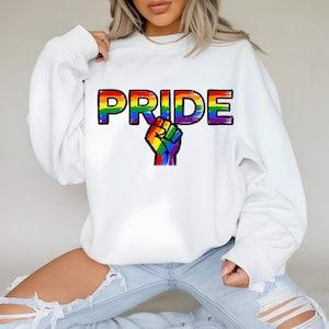 Op de afbeelding: Witte sweatshirt met het woord "PRIDE" in regenboogkleuren en een regenboogvuistafbeelding. De sweatshirt heeft een ronde hals en lange mouwen.