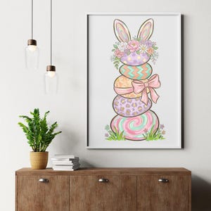 May include: A framed Easter print showing a stack of decorated eggs in pastel colours. The top egg has bunny ears and a floral crown. The eggs are decorated with patterns and a pink bow. The artwork is set against a white background.