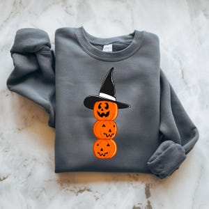 Puede incluir: Una sudadera gris oscuro con un diseño de calabazas apiladas. Las calabazas son naranjas con caras talladas y están rematadas con un sombrero de bruja negro. La sudadera está doblada sobre una superficie de mármol.