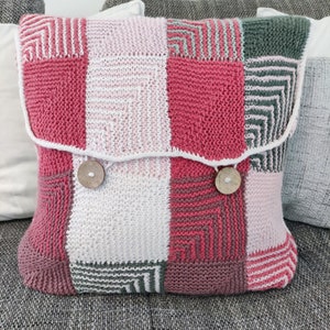 Könnte beinhalten: Ein gestricktes Kissen mit Patchwork-Muster in den Farben Rosa, Rot, Weiß und Grau. Das Kissen hat einen weißen Rand und zwei Holzknöpfe.