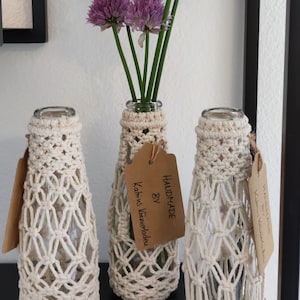 Könnte beinhalten: Drei Glasflaschen mit gehäkelten Bezügen stehen auf einem Regal. Die Flaschen sind mit lila Blumen gefüllt und die gehäkelten Bezüge sind weiß. Ein braunes Etikett ist an der mittleren Flasche befestigt mit dem Text "HANDMADE BY Kathrin Kauerlodeu".