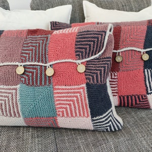 Könnte beinhalten: Zwei gestrickte Kissenbezüge mit Patchwork-Muster in Rosa-, Blau- und Weißtönen. Die Kissen haben einen weißen Rand und Holzknöpfe.