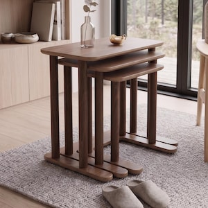 May include: A set of three nesting tables crafted from dark brown wood, featuring rounded rectangular tops and cylindrical legs. The tables are arranged in a cascading stack, with a clear glass vase containing a single stem placed on the top surface.