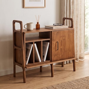 May include: A medium-toned wooden storage cabinet featuring an open top shelf, two vertical dividers for book storage, and a single cabinet door with a black metal handle. The unit has integrated side handles and stands on four tapered wooden legs.