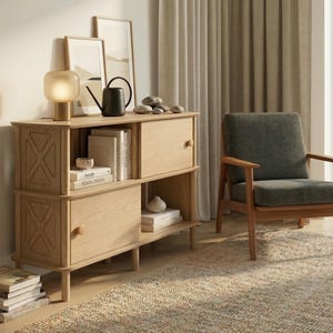 May include: A light oak cabinet with two sections, holding a lamp, books, and ornaments. A dark grey armchair with wooden arms is on a woven rug. Neutral curtains and framed art complete the interior scene.