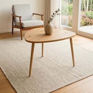 May include: A light wood, triangular coffee table with three legs, a beige rug, and a gray armchair with wooden arms. A small vase with greenery sits on the table. Two framed abstract art pieces hang on the wall.