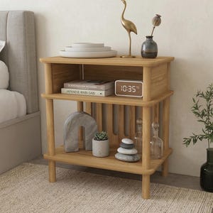 May include: A light-colored wooden nightstand with three shelves. The top shelf holds white plates, a gold bird statue, and a small vase. The middle shelf has books and a digital clock. The bottom shelf displays decorative items.