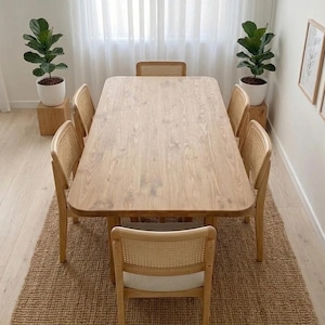 May include: A light wood dining table with six matching chairs, set on a woven jute rug. Two potted plants flank the table, and the room has a bright, airy feel. The chairs have woven backs and light-colored cushions.