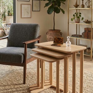 Può includere: Un set di tre tavolini ad incastro in legno chiaro con angoli arrotondati e gambe cilindriche. I tavolini sono disposti a cascata su un tappeto di iuta intrecciata. Una poltrona grigia e una scaffalatura dorata sono sullo sfondo. I tavolini misurano circa 60 cm di lunghezza.