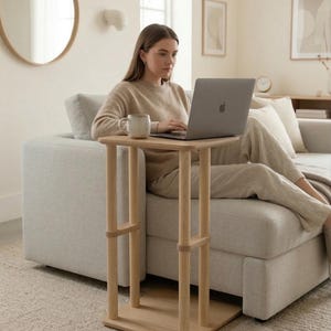 May include: A light-colored wooden side table with a square top and a lower shelf. The table has four vertical supports and is positioned next to a light gray sofa. A person is using a laptop on the table.