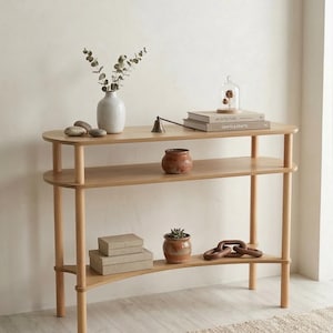 May include: A light wood, three-tiered console table with rounded edges. The top shelf holds a white vase with greenery, stones, and books. The middle shelf has a small pot. The bottom shelf has boxes, a plant, and decorative wooden links.
