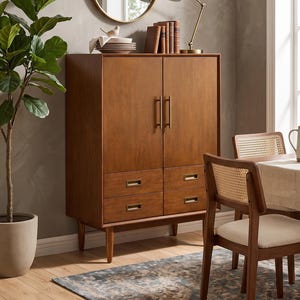 May include: A mid-century modern style wooden cabinet with two doors and four drawers. The cabinet has brass hardware and sits on tapered legs. A gold-framed mirror and a brass desk lamp are on top. A potted plant and dining chairs are also visible.