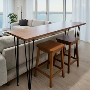Könnte beinhalten: Modernes Wohnzimmer mit hellem Sofa, einem Bartisch aus Holz mit schwarzen Haarnadelbeinen und zwei Hockern aus Holz. Große Fenster bieten einen malerischen Blick auf das Wasser und den Himmel, mit weißen Vorhängen. Ein Teppich und eine Pflanze ergänzen die Einrichtung.