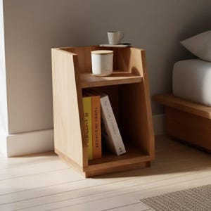 Op de afbeelding: Een lichtgekleurd houten nachtkastje met twee planken. De bovenste plank bevat een witte mok en een kleine kop met een schotel. De onderste plank toont twee boeken. Het nachtkastje staat naast een bed met wit beddengoed.