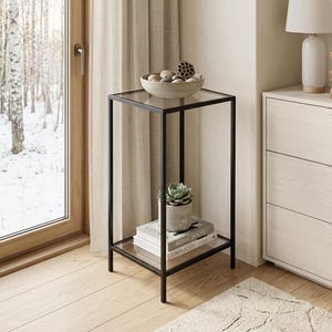 Pode incluir: Uma mesa lateral quadrada com uma estrutura de metal preto fina e duas prateleiras de vidro transparente. A prateleira superior contém uma tigela bege cheia de pedras e uma vagem de lótus seca. A prateleira inferior contém uma suculenta em vaso.