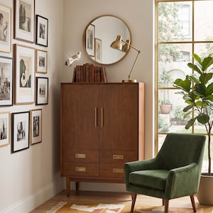 Op de afbeelding: Een moderne woonkamer met een houten kast, een groene fluwelen fauteuil en een patroonkleed. Een ronde gouden spiegel en een messing bureaulamp staan op de kast. Ingelijste kunstwerken sieren de muur en een potplant voegt groen toe.