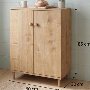 Puede incluir: Un armario de madera clara con dos puertas y pomos redondos. El armario mide 85 cm de alto, 60 cm de ancho y 30 cm de profundidad. Una taza, pan y una peque&ntilde;a planta est&aacute;n encima.