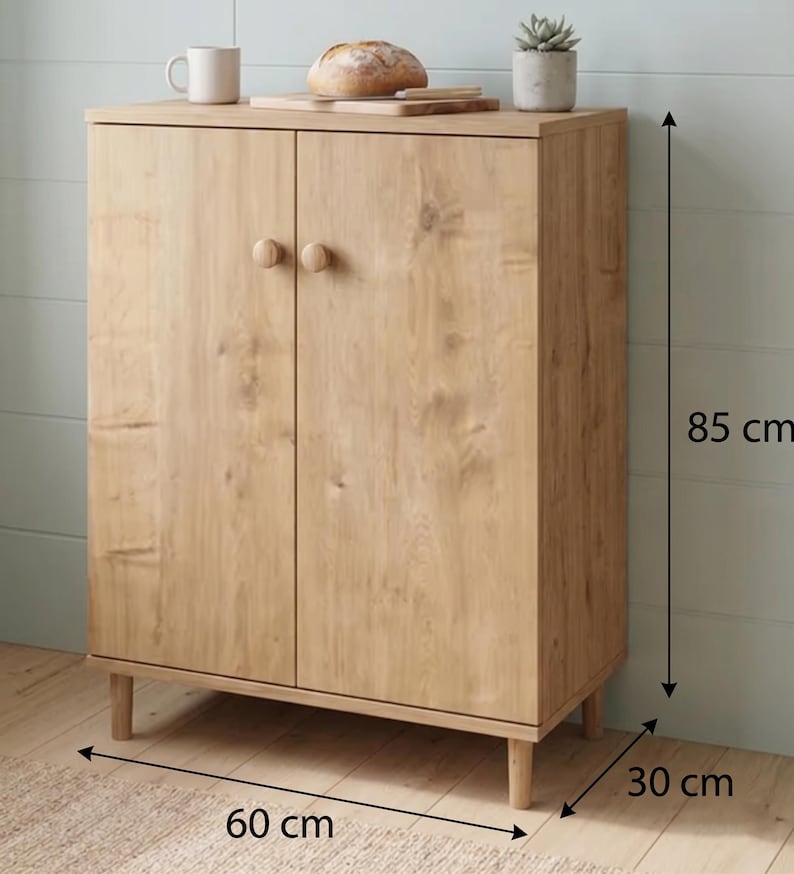 Puede incluir: Un armario de madera clara con dos puertas y pomos redondos. El armario mide 85 cm de alto, 60 cm de ancho y 30 cm de profundidad. Una taza, pan y una peque&ntilde;a planta est&aacute;n encima.