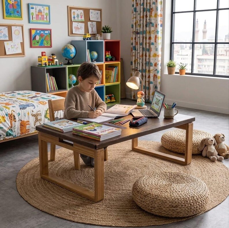 Op de afbeelding: Kinderslaapkamer met een lage houten tafel, boeken en een laptop. Een rond jute vloerkleed ligt op de vloer, met twee geweven poefs en knuffels in de buurt. Kleurrijke kunstwerken en opbergkubussen sieren de muren.
