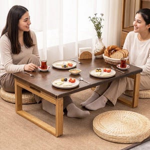 Op de afbeelding: Een lage, rechthoekige houten tafel met een donkerbruin oppervlak en lichtbruine poten. Twee mensen zitten rond de tafel, die is gedekt met borden met eten, thee en een broodmand. Geweven vloerkussens zijn zichtbaar.