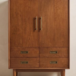 Op de afbeelding: Een houten kast in mid-century modern stijl, met twee deuren en vier laden. De kast is warmbruin met messingkleurige handgrepen en lade-trekkers. Hij staat op vier taps toelopende poten.