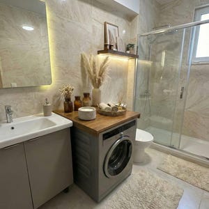 Könnte beinhalten: Modernes Badezimmer mit Waschmaschine, die in den Waschtisch integriert ist. Der Raum verfügt über eine neutrale Farbpalette mit beigen Fliesen, eine Glasdusche und dekorative Elemente wie Pampasgras und Kerzen. Ein flauschiger Teppich sorgt für Textur.