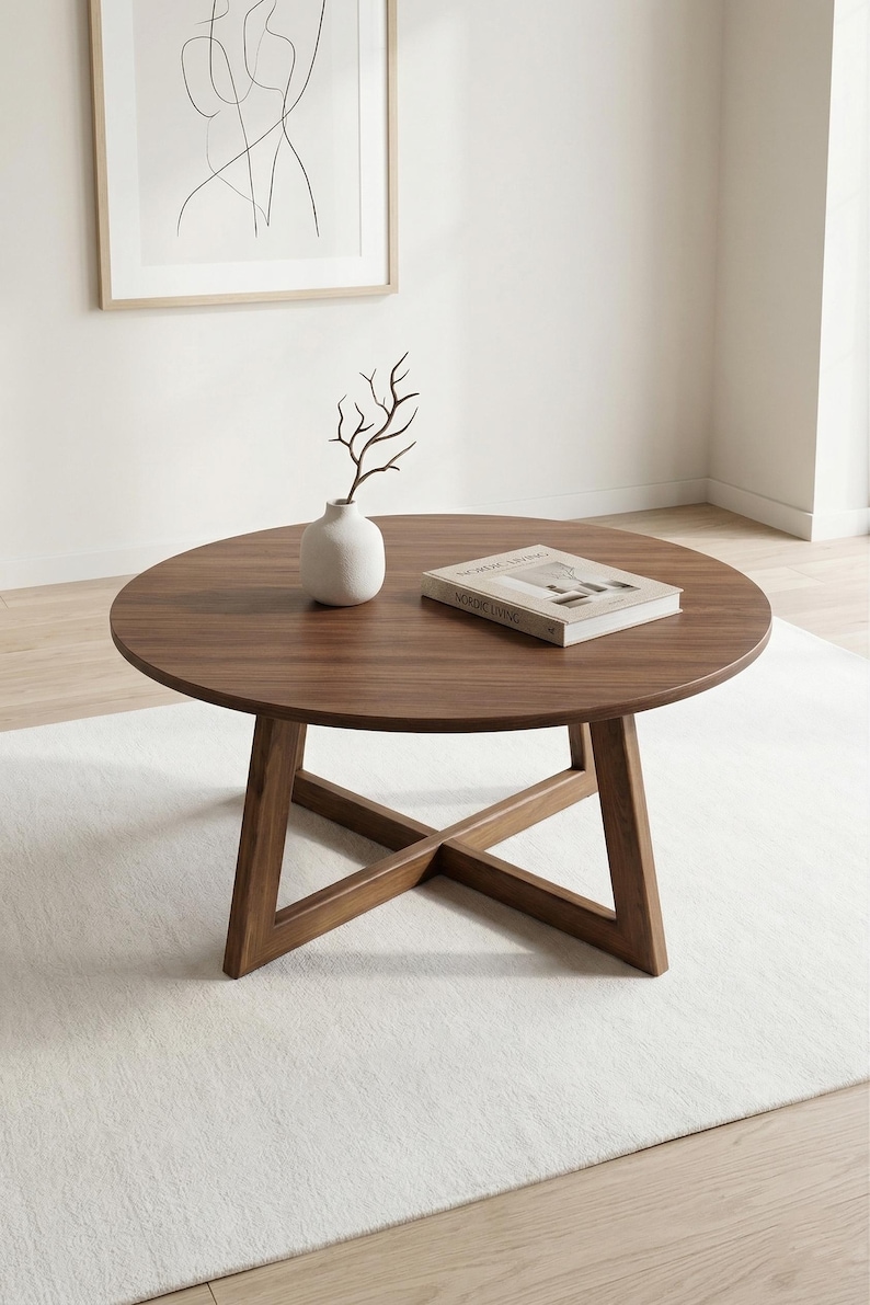 May include: A round, dark brown wooden coffee table with a cross-base design. On the table sits a small, matte white ceramic vase containing a single bare branch and a rectangular book titled Nordic Living with a white cover.