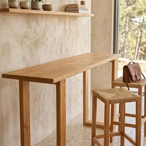 May include: A light oak bar table with a rectangular top and four square legs. Two matching bar stools with woven seats are in front of the table. The room has wooden shelves with decorative items.