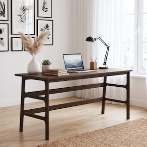 May include: A dark brown wooden desk with a rectangular top and a structural frame featuring two horizontal support bars on each side. The desk holds a silver laptop, a white vase with dried pampas grass, a small potted succulent, and a black lamp.